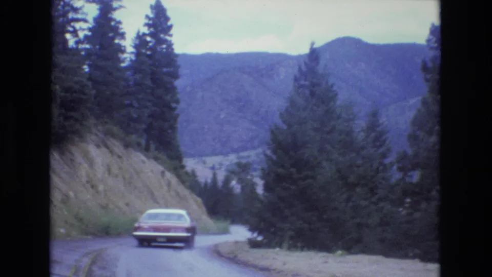 The red sedan approaches a right-hand bend on a wet Colorado mountain road, with tall evergreens, exposed roadside slope, and distant blue mountains.