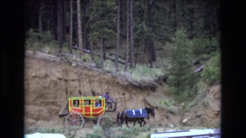 A visitor-shot 8mm frame shows the red and yellow carriage and dark horse below tall trees, with the road and hillside partly cropped by the film frame.