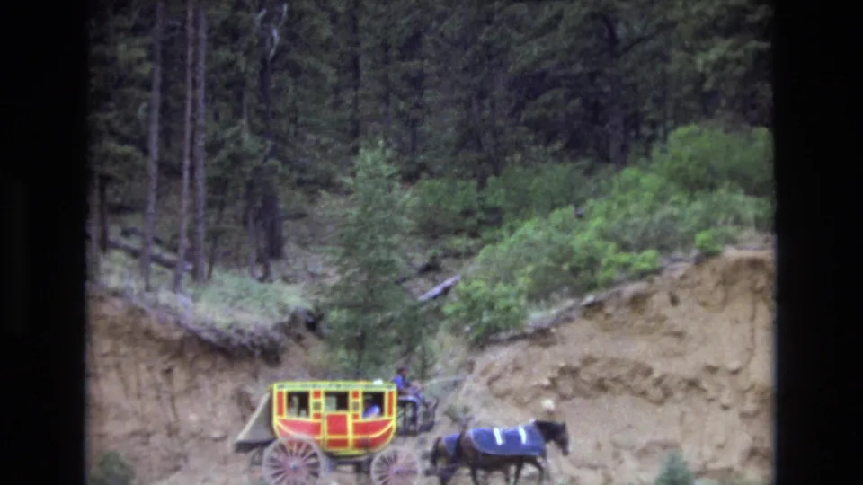 The carriage and horses continue along the mountain road, with a steep exposed dirt bank and tall trees behind them in the Colorado home-movie footage.
