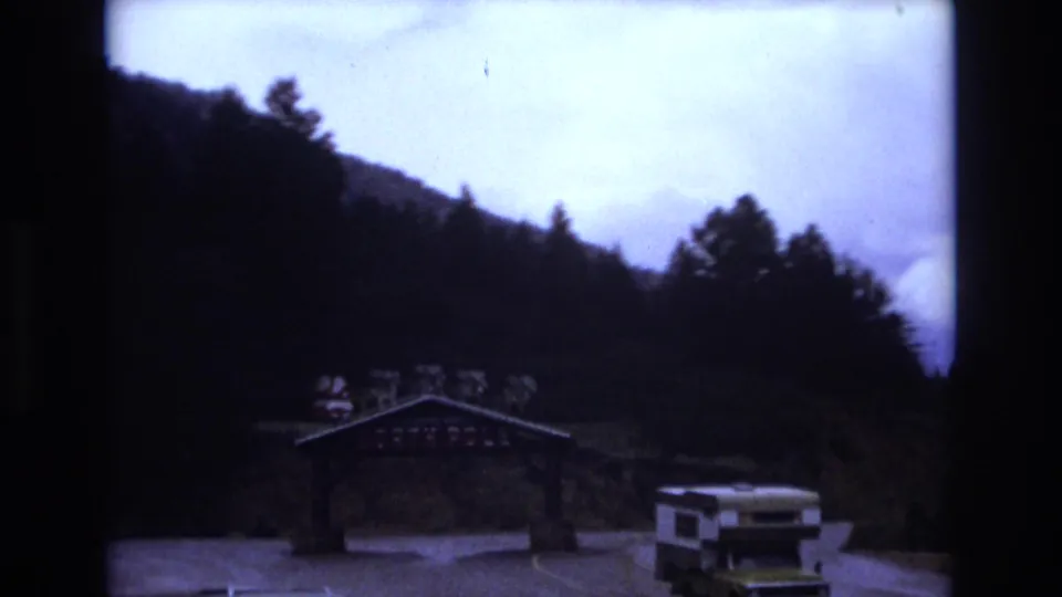 A wooden roadside entrance arch decorated with small Santa figures stands below forested hills, with a vintage camper van nearby in 1973 Colorado footage.