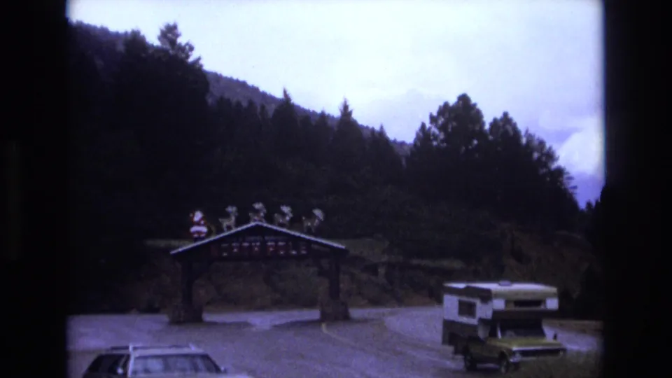 The roadside archway with Santa figures is seen from farther back, with a camper van and car at the entrance against wooded Colorado hills.