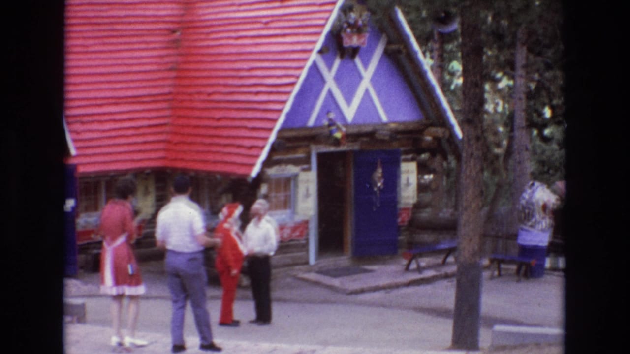 1973 Colorado 8mm home-movie frame showing adult visitors near a Santa-costumed figure outside a decorative log cabin with a red roof and purple facade.