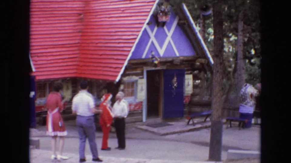 1973 Colorado 8mm home-movie frame showing adult visitors near a Santa-costumed figure outside a decorative log cabin with a red roof and purple facade.