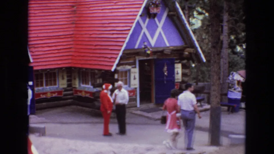 1973 Colorado home-movie frame of a Santa-costumed figure conversing with an adult man near a Christmas-themed log cabin while other visitors walk through the area.