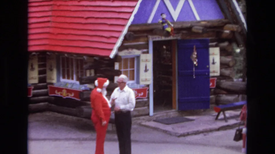 Closer 1973 Colorado 8mm frame showing a Santa-costumed figure and an adult man in conversation beside the open doorway of a red-roofed Christmas village cabin.