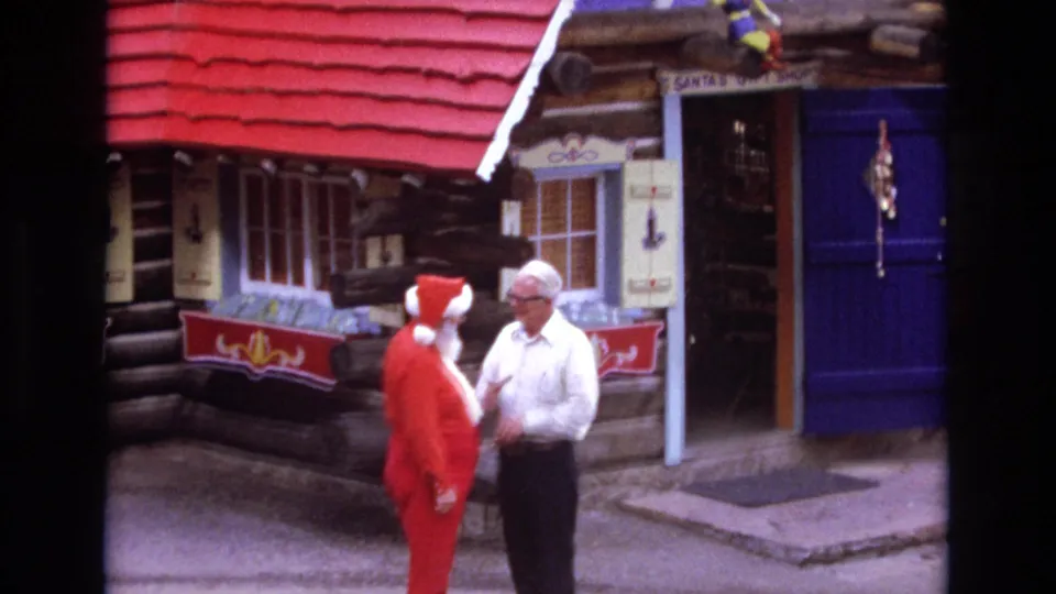 1973 Colorado home-movie frame with a Santa-costumed figure and an adult man standing outside a log-cabin Christmas village storefront with blue door panels and visible signage.