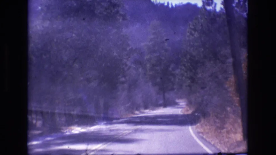 1973 Colorado home-movie frame from a moving vehicle, looking ahead at a quiet paved road curving through a forested mountain area.