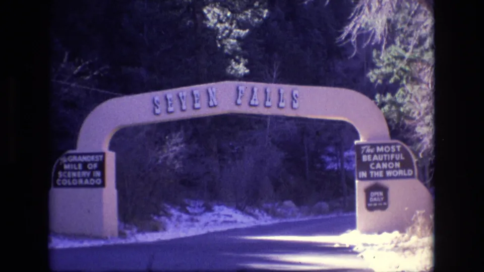 1973 Colorado home-movie frame showing the Seven Falls arch spanning the road, with signs reading Grandest Mile of Scenery in Colorado and The Most Beautiful Canon in the World.