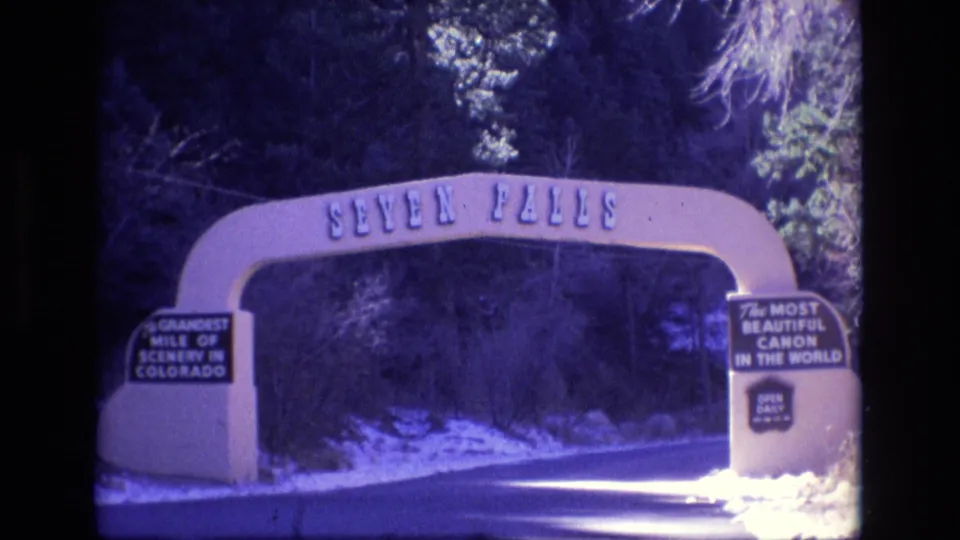 1973 Colorado home-movie frame with the Seven Falls arch spanning the entrance road, visible slogan signs, winter snow patches, and a wooded canyon setting.