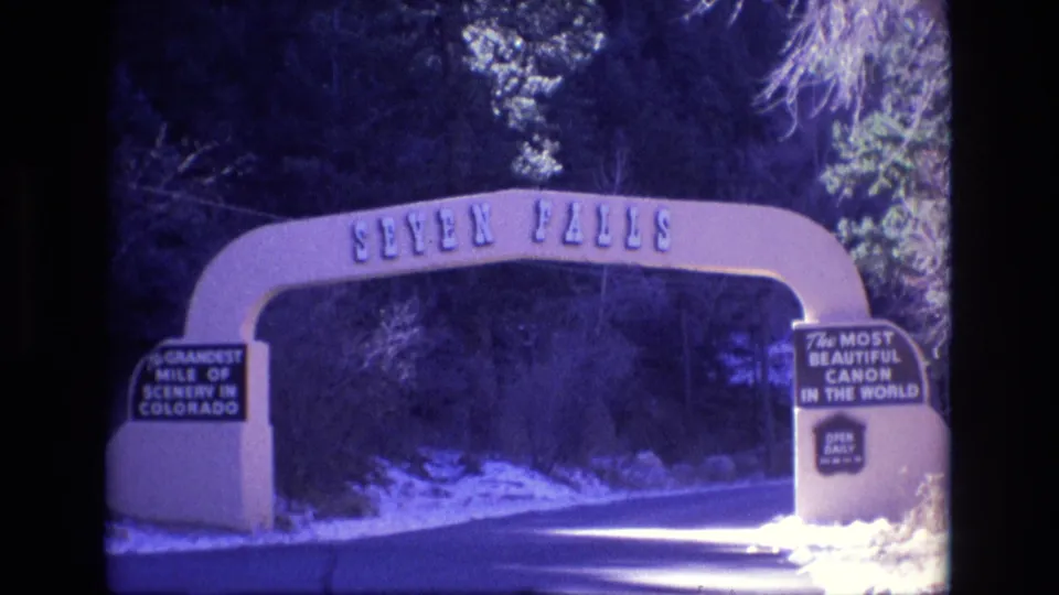 1973 Colorado 8mm frame holding on the Seven Falls entrance arch, with the paved road, snowy shoulders, dark trees, and attraction signs still visible.