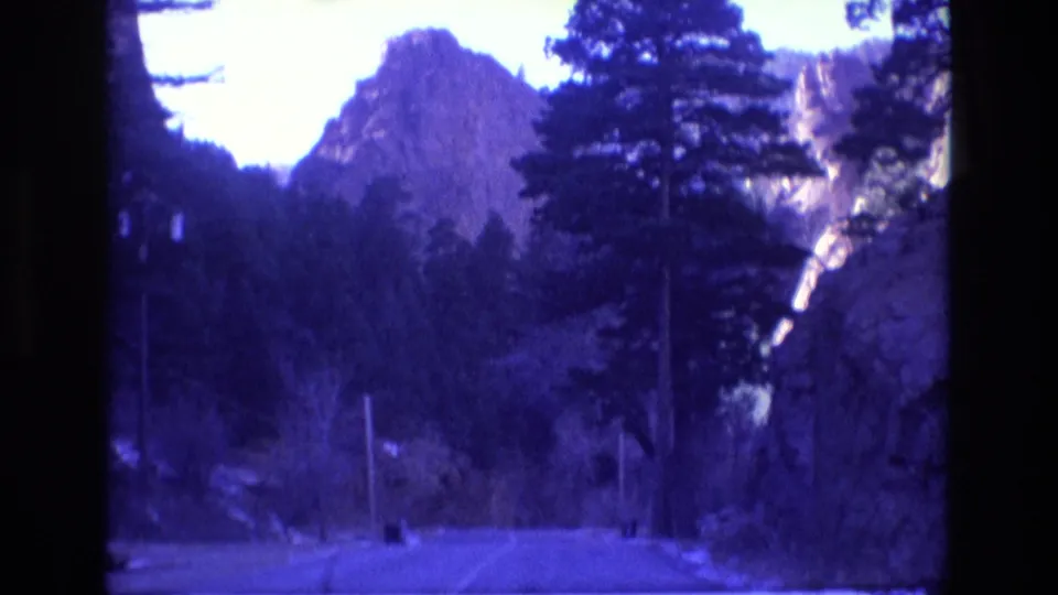 1973 Colorado home-movie frame showing a paved mountain road receding through pine trees toward rocky canyon formations, with the same aged purple-blue film cast.