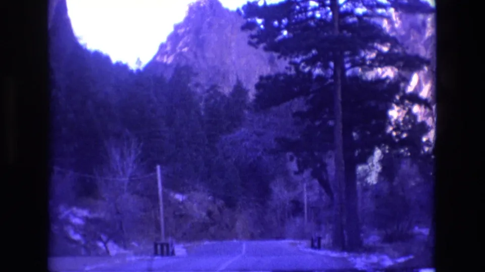 1973 Colorado home-movie frame looking along a quiet paved road bordered by pine trees, utility poles, and steep rocky mountain terrain.