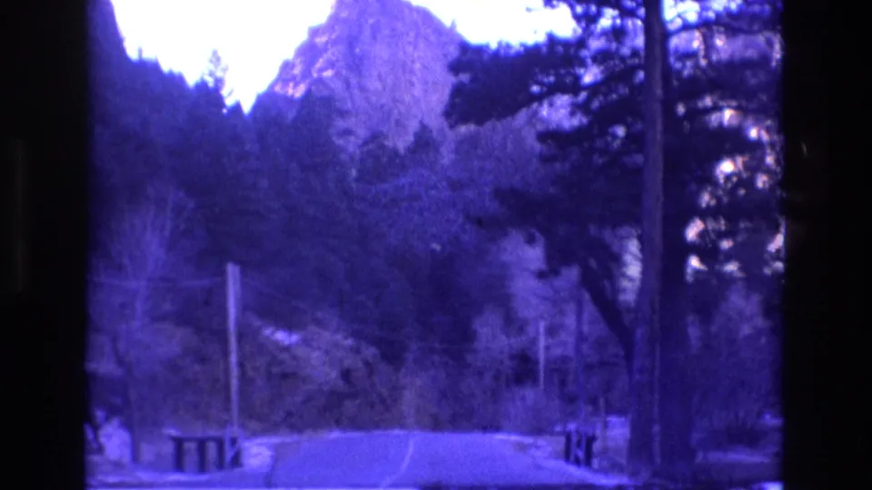 1973 Colorado 8mm frame of a curving mountain road with pine trees, roadside railings, utility lines, and small patches of snow near the forest edge.