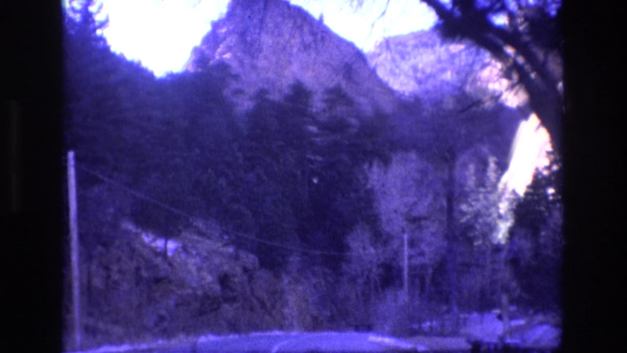 1973 Colorado 8mm home-movie frame showing a road through dark trees toward steep mountain and rock formations, with a purple-blue film cast.