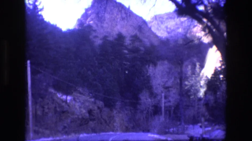 1973 Colorado 8mm home-movie frame showing a road through dark trees toward steep mountain and rock formations, with a purple-blue film cast.