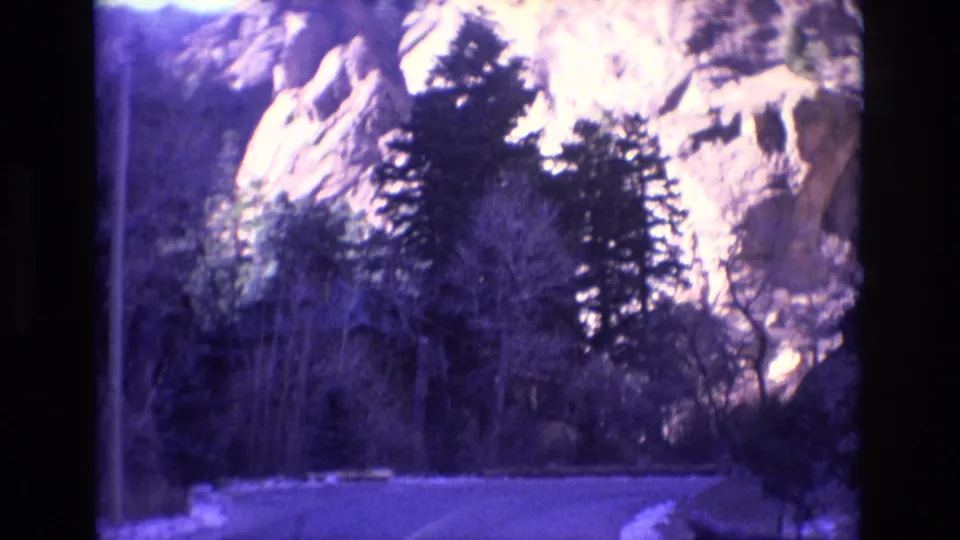 1973 Colorado 8mm frame showing a curving mountain road below pale rock cliffs, with evergreens and bare roadside trees visible through home-movie grain.