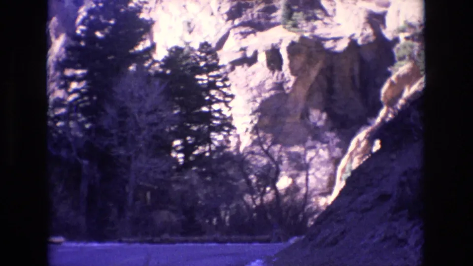 1973 Colorado home-movie frame from a vehicle passing a roadside rock slope, with evergreens, bare trees, and sunlit cliffs beyond.