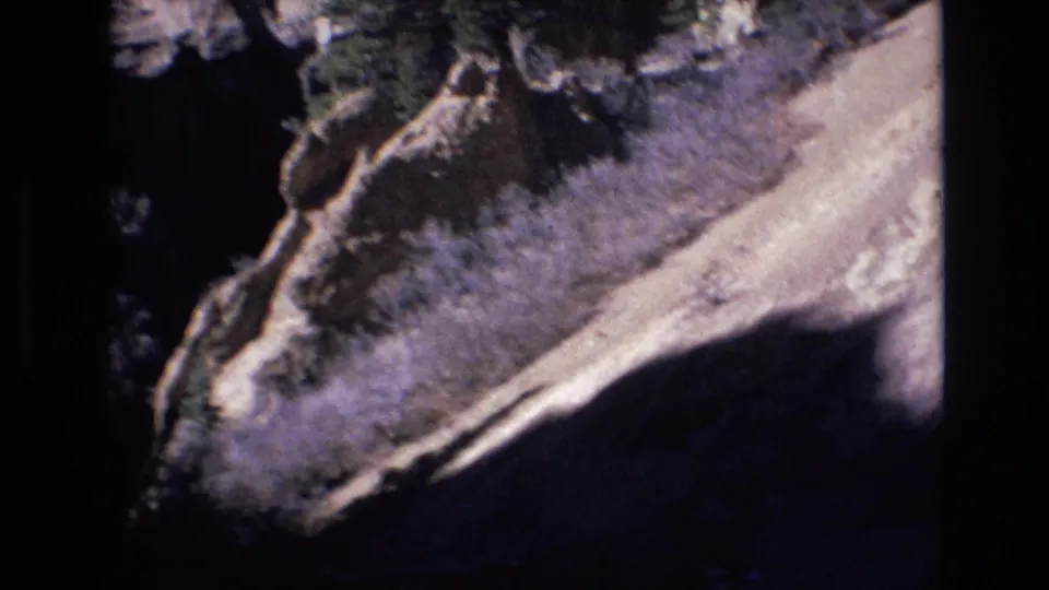 1973 Colorado 8mm frame looking along a curving mountain road bordered by rough rock, brush, and shadowed roadside terrain.