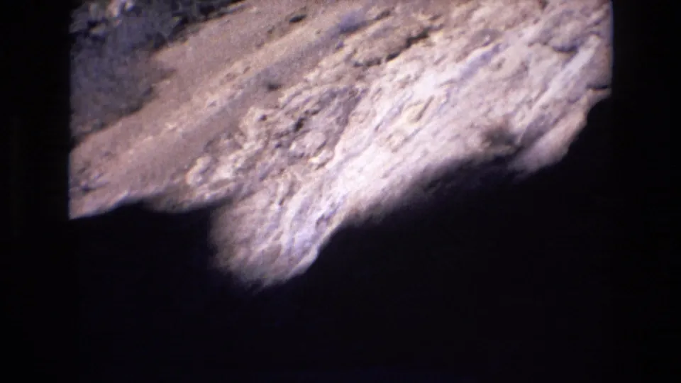 1973 Colorado 8mm frame showing a close sunlit rock slope beside the mountain road, with dark shadow at the lower edge of the frame.