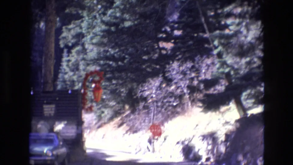 1973 Colorado home-movie footage shows a closer car-window view of the roadside booth, red stop sign, blue parked car, and wooded slope beside the road.