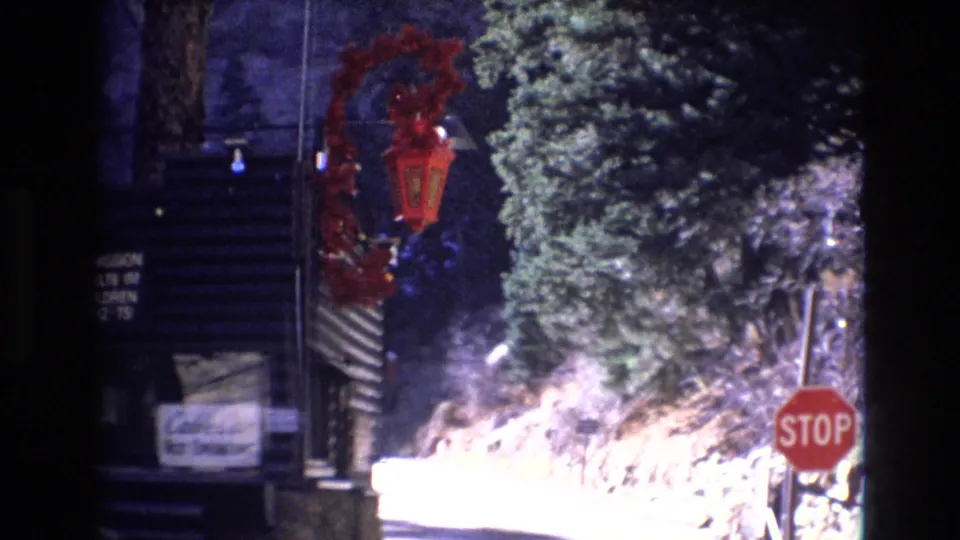 1973 Colorado 8mm frame showing a red stop sign, evergreen trees, rocky roadside terrain, and a partially visible holiday display near a rustic mountain setting.