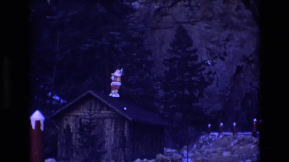 1973 Colorado 8mm frame showing a rustic wooden cabin among evergreens and rocks, with a Santa Claus figure mounted above the roofline.