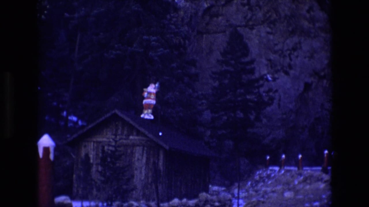 1973 Colorado 8mm frame showing a rustic cabin at night with a Santa figure on the roof, snow patches, evergreens, and a rocky cliff behind it.
