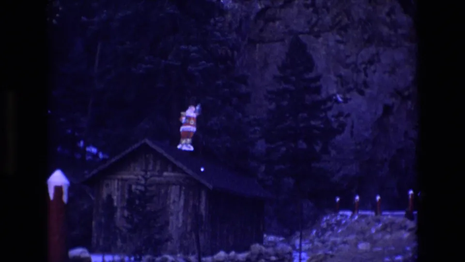 1973 Colorado 8mm frame showing a rustic cabin at night with a Santa figure on the roof, snow patches, evergreens, and a rocky cliff behind it.