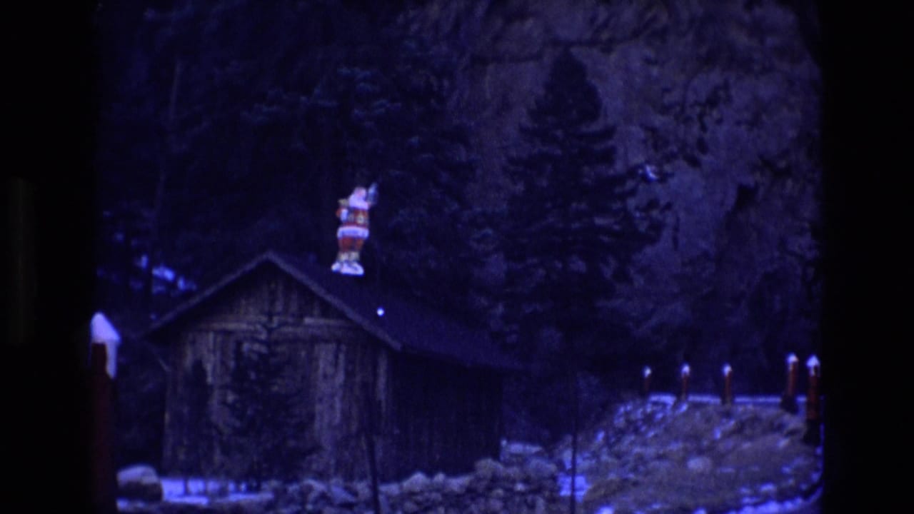 Cabin with a rooftop Santa figure, roadside posts, snow, trees, and a rocky cliff at night.