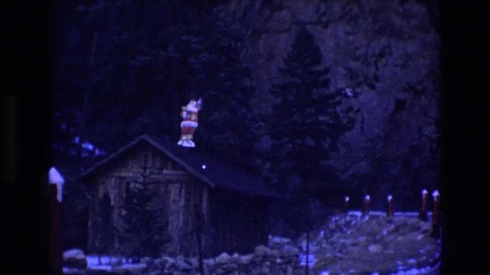 1973 Colorado home-movie frame of a dark cabin beside snow and roadside posts, with a Santa figure on the roof against trees and a cliff face.
