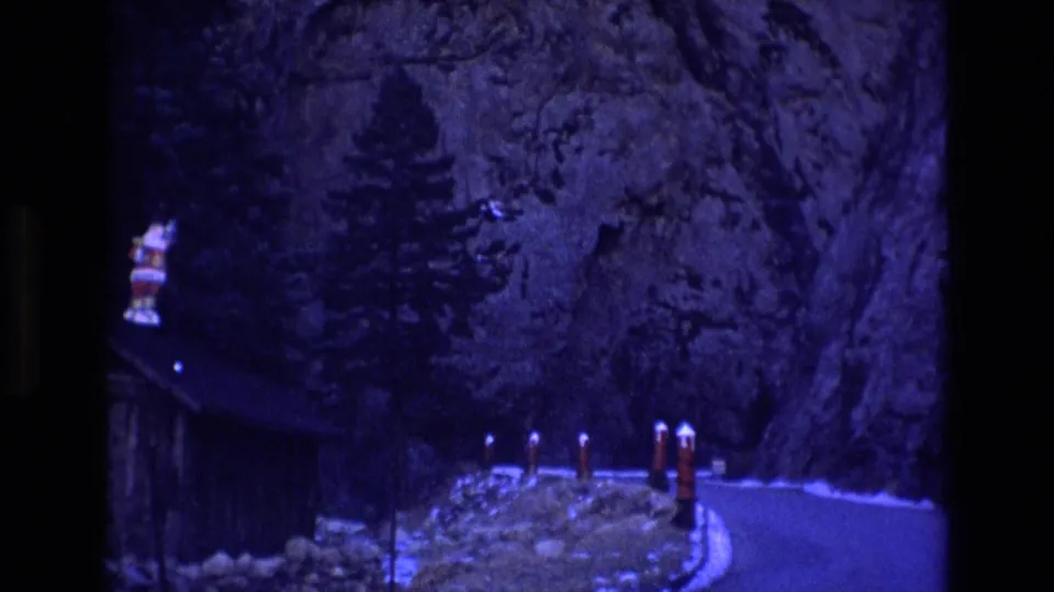 1973 Colorado 8mm frame with the cabin partly at the left edge, a Santa figure near the roofline, red-and-white posts, snow patches, a road, and a rocky mountain wall.