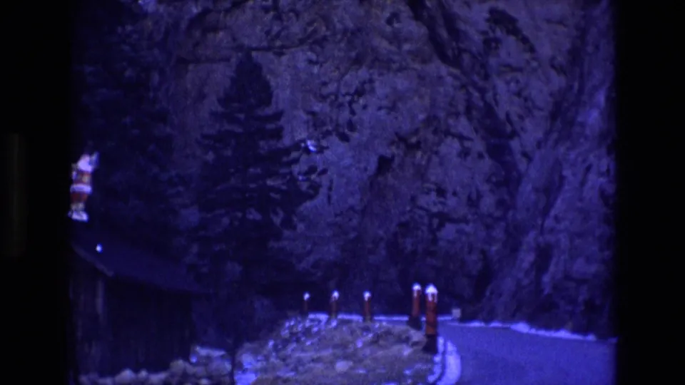 1973 Colorado frame showing the decorated cabin slipping toward the left edge while roadside posts, snow, trees, and the rocky cliff remain visible in low blue night exposure.