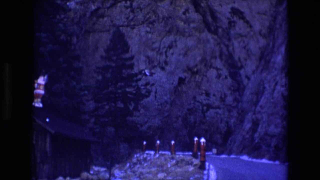 1973 Colorado 8mm frame showing a narrow mountain road below steep rock cliffs, evergreen trees, a dark structure at left, and red-and-white bollards along the roadside.