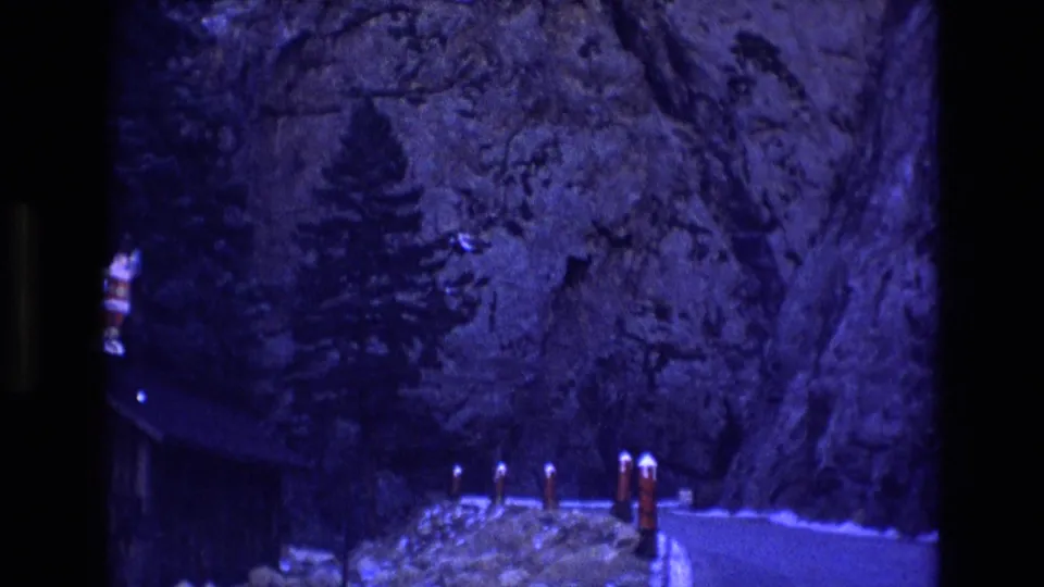 1973 Colorado home-movie frame of a blue-tinted mountain road bordered by rock walls, evergreen trees, red-and-white bollards, and a dark cabin-like form at left.