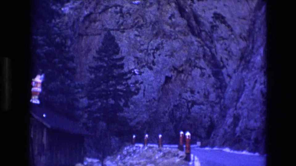 1973 Colorado 8mm frame showing a cliff-lined road with evergreen trees, red-and-white bollards, a dark building edge, and small patches of snow.