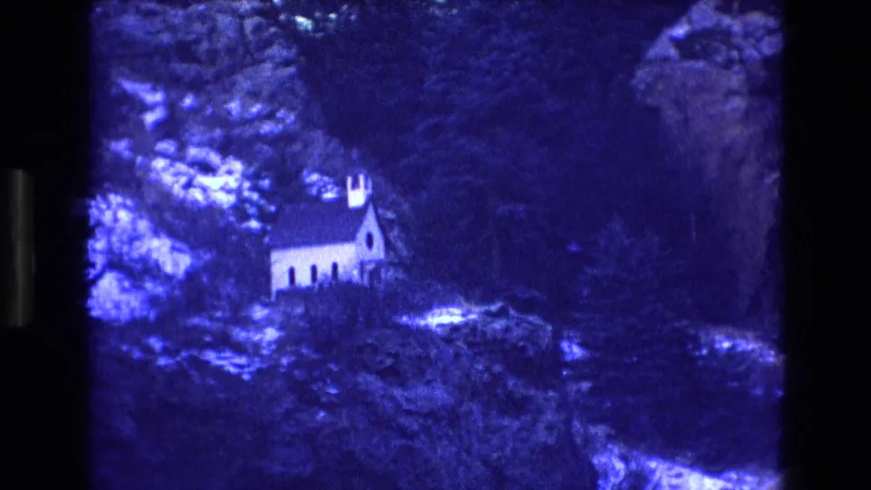 1973 Colorado 8mm frame showing a distant white church on a hillside surrounded by evergreen trees, rocky ground, and snow-dusted terrain.