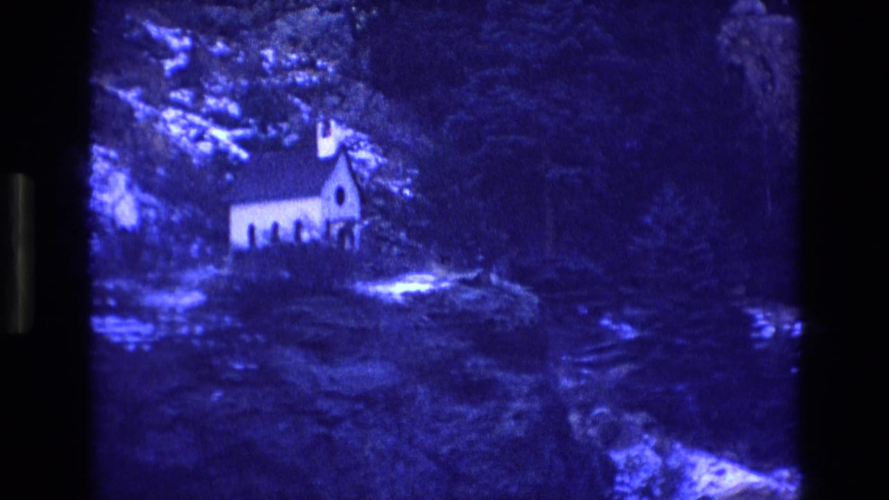 1973 Colorado 8mm frame showing a small white church set among dense trees and rocky mountain terrain under a strong blue cast.