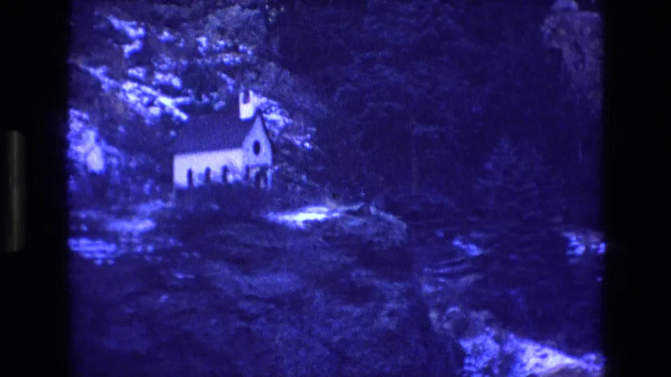 1973 Colorado 8mm frame showing a small white church set among dense trees and rocky mountain terrain under a strong blue cast.