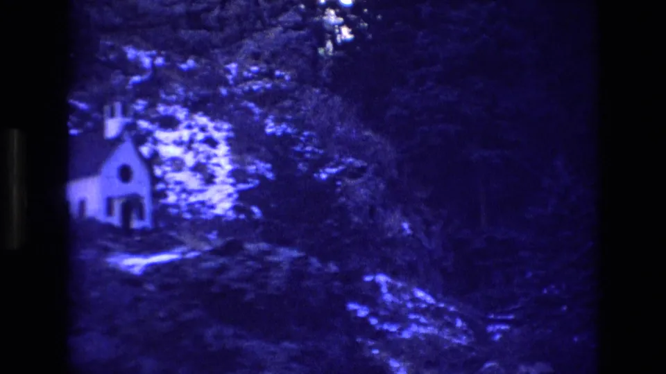 1973 Colorado home-movie frame with a small white church or chapel surrounded by trees, rocks, and high-contrast blue-tinted terrain.