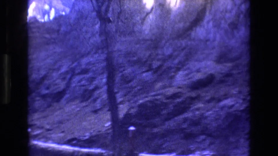 1973 Colorado travel frame showing a blue-tinted rocky slope with trees, shadowed terrain, and a pale strip of ground at the bottom of the frame.