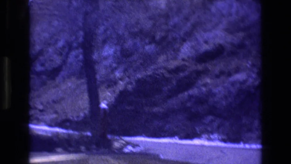 1973 Colorado 8mm frame showing a paved mountain road curving past snow, evergreen trees, and steep rock terrain with a strong purple color cast.