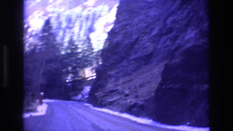 1973 Colorado 8mm frame showing a road through a rocky forested mountain area, with snow at the roadside and bright distant terrain beyond.