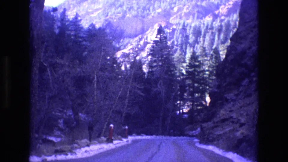 1973 Colorado 8mm frame looking down a mountain road between dark rock walls, evergreen trees, roadside snow, and distant snowy slopes.