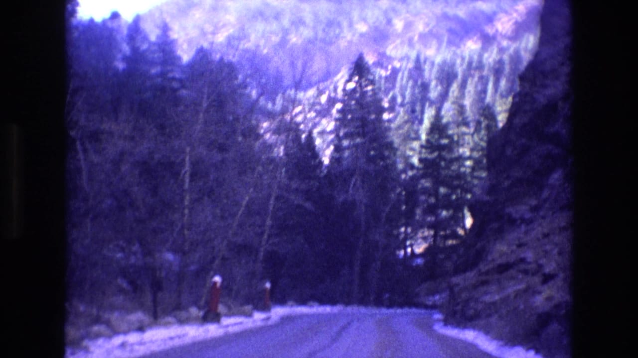 Curving snowy mountain road bordered by evergreens and a rocky cliff.