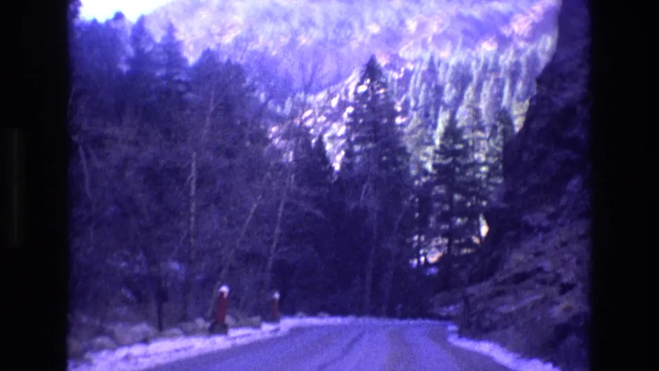1973 Colorado 8mm frame of a winding mountain road with snow at the roadside, evergreen trees, bare branches, and a dark rock wall at the right under a purple-blue color cast.