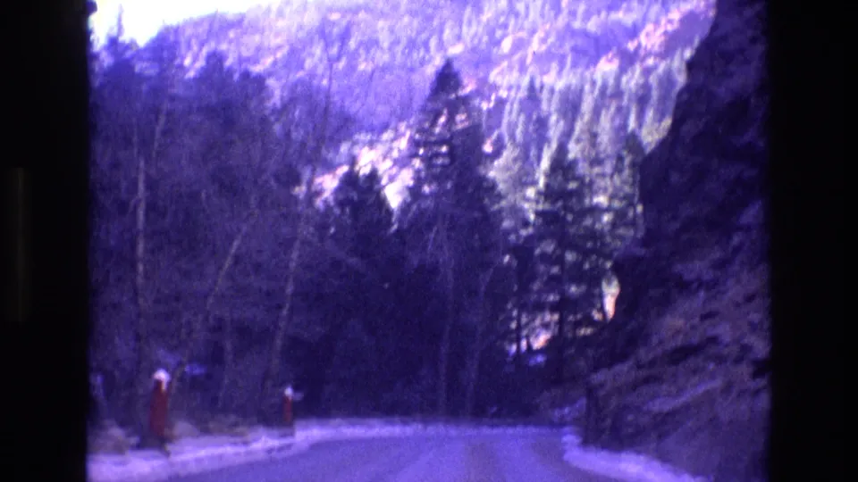 1973 Colorado home-movie frame looking along a winter mountain road as it bends through evergreens, bare trees, snow patches, and rock faces with strong blue-purple film tones.