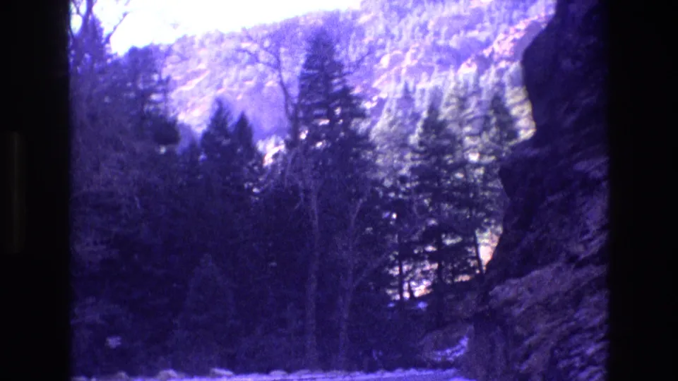 1973 Colorado archival 8mm frame showing the mountain road environment from close range, with snow at the bottom edge, dark conifers, leafless trees, a rock wall, and a washed purple-blue cast.