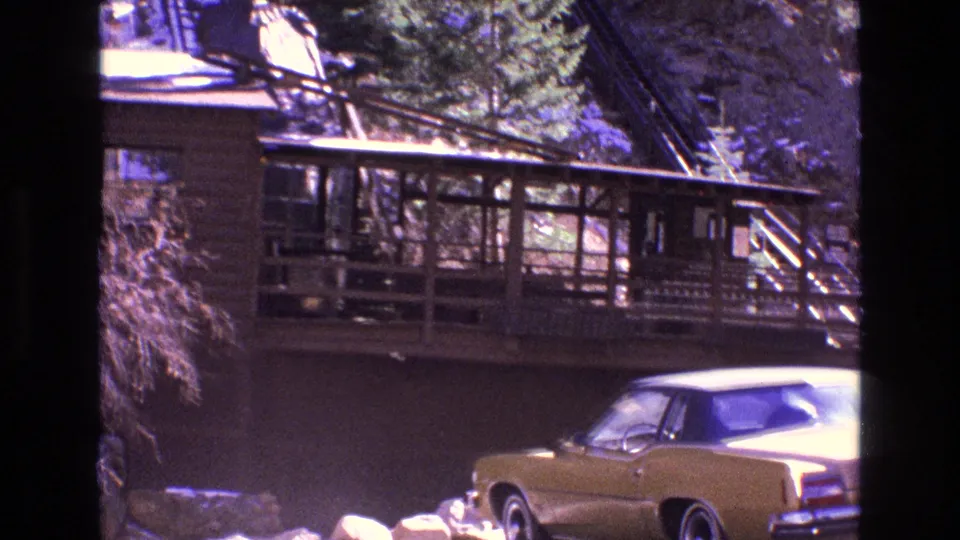 Rustic wooden bridge or station-like structure fills the frame, with a gold 1970s sedan partly visible in the lower right of the Colorado mountain scene.