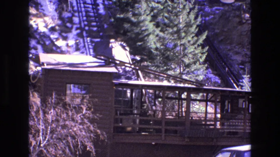 1973 Colorado home-movie frame with a wood-sided lodge, tall evergreen, snow-dusted slope, and visible inclined railway track behind the building.