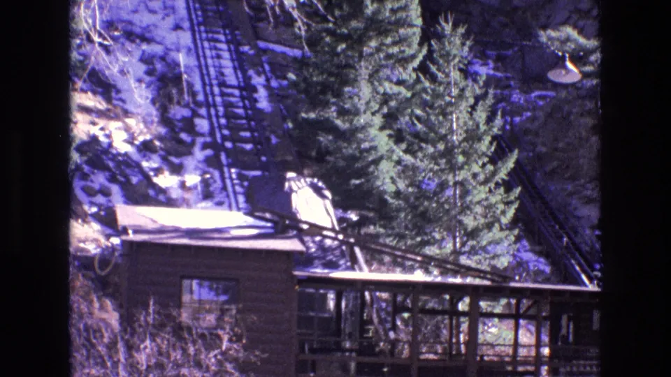 1973 Colorado 8mm frame showing the roofline and porch of a rustic lodge beneath tall evergreens, with a steep snow-lined railway track rising up the hillside.